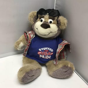 Puede incluir: Un oso de peluche con gafas de sol, una camiseta azul con el texto "STUFFED WITH USA PRIDE" y una camisa a cuadros roja y negra. El oso tiene pelaje marrón y está sentado.