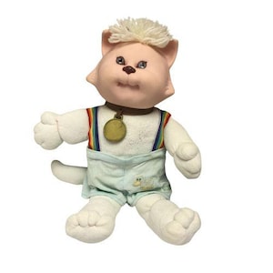 May include: A vintage plush cat doll with a light pink face and white body. The doll wears a gold medallion, rainbow suspenders, and light blue shorts. The cat has a tuft of white hair and is in a sitting position.