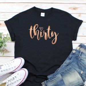 Może przedstawiać: Czarny t-shirt ze słowem "thirty" w miedzianym skrypcie, z małym sercem nad "i". Koszulka jest połączona z niebieskimi dżinsami i białymi trampkami.