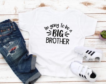 May include: White t-shirt with black text that reads "I'm going to be a BIG BROTHER". The shirt is on a wooden surface with a pair of blue jeans and white sneakers.