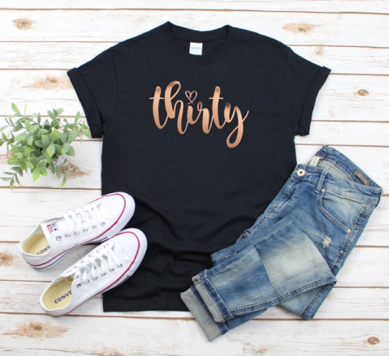 Może przedstawiać: Czarny t-shirt ze słowem "thirty" w miedzianym skrypcie, z małym sercem nad "i". Koszulka jest połączona z niebieskimi dżinsami i białymi trampkami.