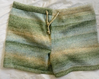 May include: Knitted shorts featuring a horizontal gradient pattern in shades of sage green, pale blue, and soft beige. The garment includes a ribbed elastic waistband and a matching knitted drawstring cord centred at the front waistline.
