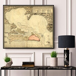 May include: A vintage map of a coastal area with detailed markings and text. The map is framed in black and displayed on a white wall above a black console table with a lamp and plants.
