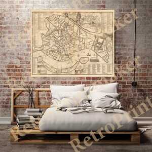 May include: An antique map of Venice, Italy, printed on aged paper with a brown and cream color scheme. The map shows the canals and streets of the city, with labels in Italian. The map is framed and hanging on a brick wall above a bed with white linens.