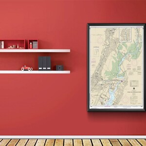 May include: A framed nautical chart of the Passaic River and Newark Bay, New Jersey. The chart is in black and white with blue and green accents. The chart is framed in a black frame and is hanging on a red wall.