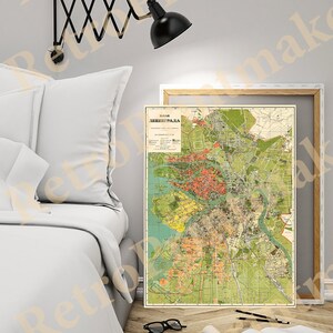May include: A vintage map of Leningrad, now known as Saint Petersburg, Russia. The map is printed in a muted color palette with shades of green, yellow, and brown. The map is framed and hanging on a wall in a room with a white bed and a black wall lamp.