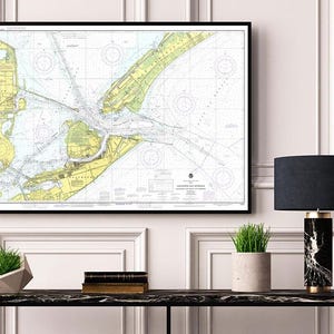 Puede incluir: Un mapa náutico enmarcado en un marco negro cuelga de una pared blanca. El mapa presenta líneas y marcas detalladas en azul, amarillo y blanco. Una lámpara negra con base de mármol y una pequeña planta en maceta están sobre una mesa negra.