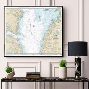 Puede incluir: Un mapa náutico enmarcado de la Bahía de Chesapeake, con marcas detalladas y un marco negro. El mapa se muestra en una pared blanca sobre una mesa consola negra con una lámpara negra, una planta en maceta y una pila de libros.