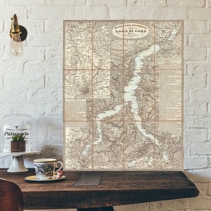 May include: An antique map of Lake Como, Italy, displayed on a wooden surface. The map is divided into grid sections and features detailed geographical information. The map is in shades of brown and beige, with text in Italian. A light bulb and a glass cloche with a plant are visible.