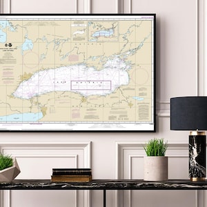 May include: A framed nautical chart of Lake Ontario, with a black frame and a beige background. The chart shows the lake's shoreline, islands, and depths. The title "Lake Ontario" is printed in the center of the chart. The chart is hanging on a white wall above a black console table with a lamp, a potted plant, and a stack of books.