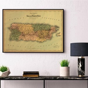May include: Vintage map of Puerto Rico, framed in black, with a green, yellow, and pink color scheme. The map is hanging on a white wall above a black table with a lamp and plants.