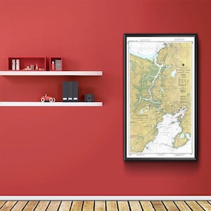 May include: A framed nautical chart of a coastal area, featuring detailed depictions of waterways, landmasses, and navigational information. The chart is in a black frame and hangs on a red wall. Shelves with decorative items are below.