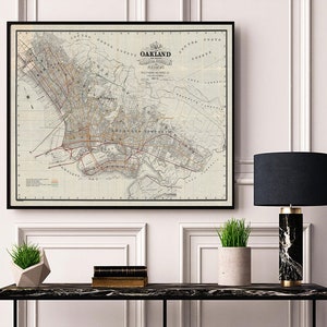 May include: A vintage map of Oakland, California, framed in black, hangs above a black marble console table with a black and white marble lamp and a potted plant. The map is dated 1892 and shows the city's streets and landmarks.