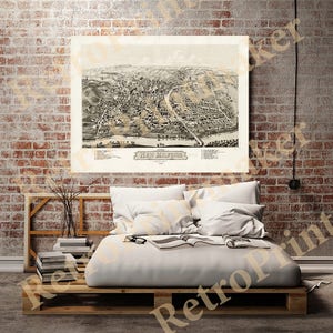 May include: A vintage black and white map of New Milford, framed and hanging on a brick wall. The map is above a bed made from wooden pallets, with white pillows and a blanket. A stack of books and a vase with branches are on the side.