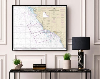 May include: A framed nautical chart of the San Diego to San Francisco Bay area, with purple lines marking shipping lanes. The chart is hanging on a white wall above a black console table with a lamp, a potted plant, and a stack of books.