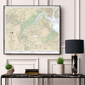 May include: A framed nautical chart of the Raritan River, New Jersey, with detailed markings and a beige background. The chart is hanging on a white wall above a black console table with a lamp, a potted plant, and a stack of books.