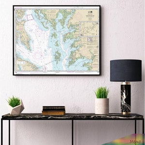 May include: A framed nautical chart of the Chesapeake Bay, with detailed markings and a smaller inset map. The chart is hanging on a white wall above a black console table with a lamp and potted plants.