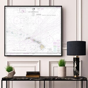 May include: A framed nautical chart of the Hawaiian Islands, showing the islands and surrounding waters. The chart is in black and white with some colored lines and text. The chart is hanging on a white wall above a black console table with a lamp, plants, and books.
