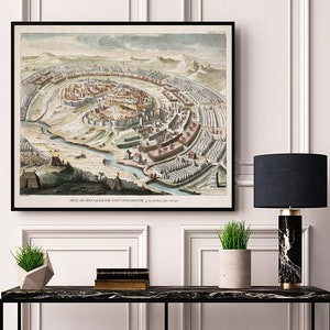 May include: A vintage illustration of Jerusalem, showing the city surrounded by a wall and a moat. The illustration is in a black frame and is hanging on a white wall. There is a black lamp and a potted plant on a black table in front of the illustration.