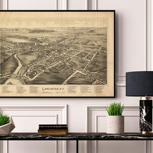May include: A vintage black and white map of Lancaster, NY, framed in a black frame and hanging on a white wall above a black console table with a black and white marble lamp, a potted plant, and a stack of books.