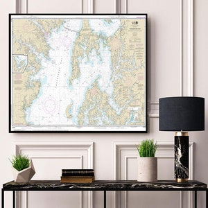 May include: A black framed nautical chart of Chesapeake Bay hangs on a white wall above a black console table. The table has a black lamp with a dark gray shade, a potted plant, and a stack of books on it.