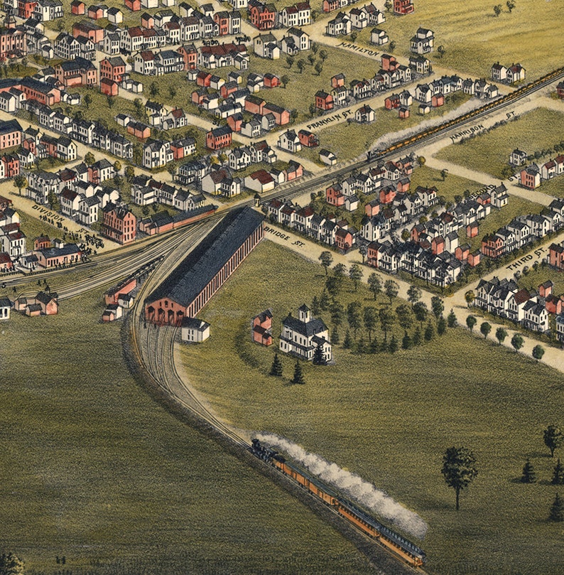 May include: An aerial view of a train station with a large train shed and multiple tracks. The station is surrounded by a residential neighborhood with houses and trees. A train is pulling out of the station with smoke billowing from the engine.