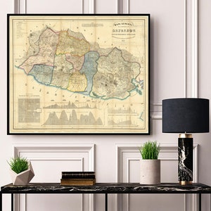 Op de afbeelding: Een vintage kaart van de regio "Santa Marta" in Colombia, gedrukt in 1853. De kaart is zwart ingelijst en toont een gedetailleerde omtrek van de regio met verschillende kleuren en markeringen. De kaart wordt aan een muur getoond boven een zwarte consoletafel met een lamp en planten.