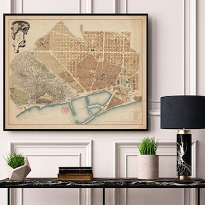 May include: A vintage map of Barcelona, Spain, printed in black and white with red and blue accents. The map shows the city's streets, buildings, and landmarks. The map is framed in a black frame and is hanging on a white wall.