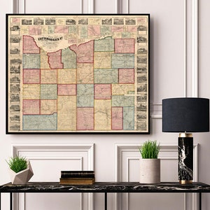 Pode incluir: Um mapa vintage do condado de Cattaraugus, Nova Iorque, impresso em preto, vermelho, verde e azul. O mapa está emoldurado e pendurado em uma parede branca acima de uma mesa preta com uma lâmpada e plantas.