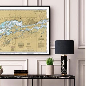 May include: A framed nautical chart with a black border, depicting a detailed map of a coastal area. The chart features blue and tan tones, with intricate lines and symbols. The image is displayed above a black marble table, with a black lamp and small potted plants.