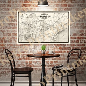 Op de afbeelding: Een vintage kaart van de La Crosse and Milwaukee Rail Road, gedrukt in zwart-wit, hangt aan een bakstenen muur boven een kleine ronde tafel met twee stoelen. De tafel heeft een houten blad en een zwart metalen onderstel. Er staat een potplant op de tafel, samen met twee wijnglazen en een kopje koffie.