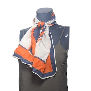 May include: A lightweight scarf featuring a geometric pattern with bold blocks of white, navy blue, and bright orange. The scarf is tied in a simple knot around the neck of a dark gray mannequin torso wearing a dark gray camisole top.