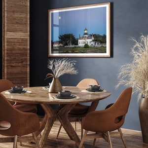 May include: A round wooden dining table with four brown leather chairs. The table is set with four place settings, each with a black plate, a black bowl, and a silver fork and knife. A framed photograph of a lighthouse on a beach hangs on the wall behind the table.