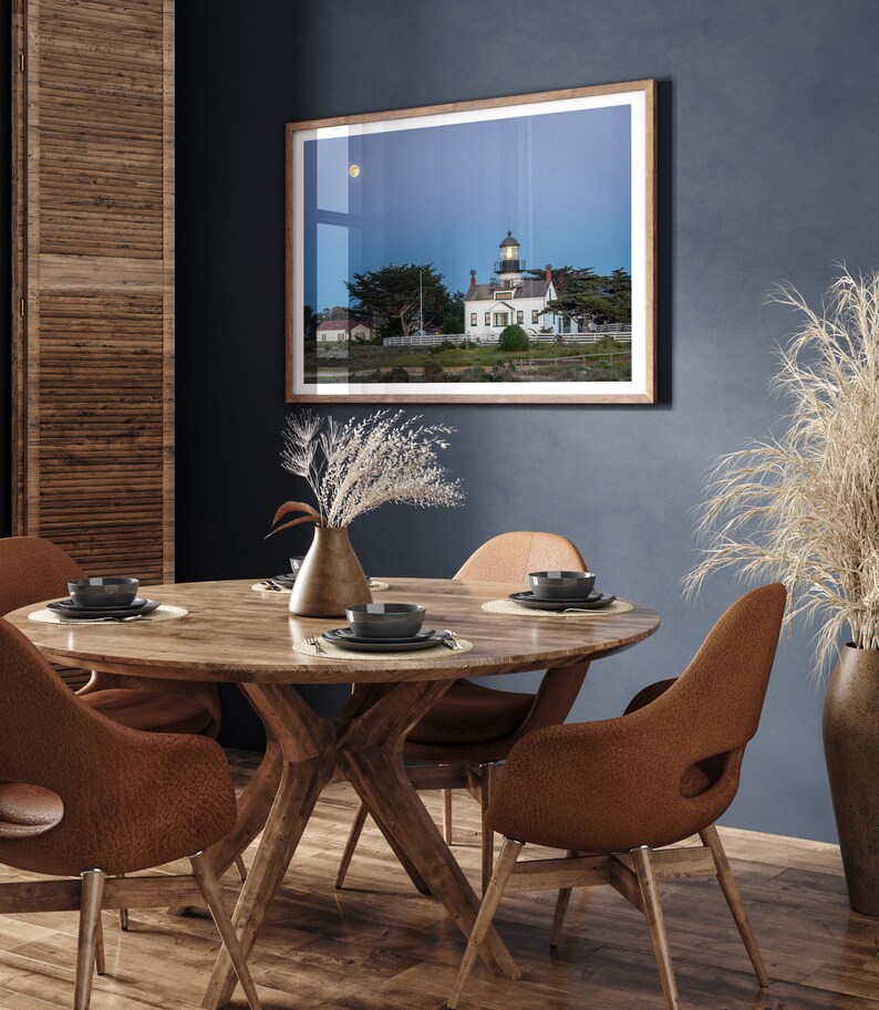 May include: A round wooden dining table with four brown leather chairs. The table is set with four place settings, each with a black plate, a black bowl, and a silver fork and knife. A framed photograph of a lighthouse on a beach hangs on the wall behind the table.