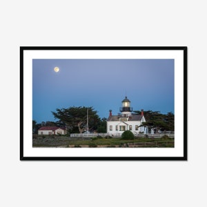 May include: A white lighthouse with a green roof and a lit window stands on a grassy hill under a blue sky with a full moon.  The lighthouse is surrounded by trees and a white fence.