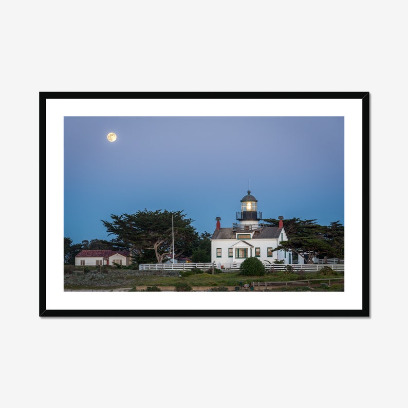 May include: A white lighthouse with a green roof and a lit window stands on a grassy hill under a blue sky with a full moon.  The lighthouse is surrounded by trees and a white fence.