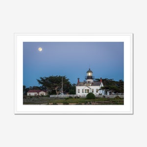 May include: A white lighthouse with a green roof stands on a grassy hill with trees in the foreground. The moon is visible in the blue sky.