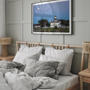 May include: A white lighthouse with a black roof and a window with a view of the moon. The lighthouse is surrounded by trees and grass. The image is framed and hanging on a wall in a bedroom with a bed and bedside tables.