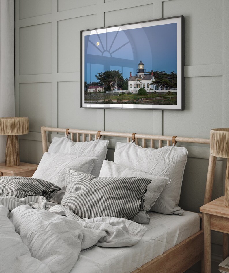 May include: A white lighthouse with a black roof and a window with a view of the moon. The lighthouse is surrounded by trees and grass. The image is framed and hanging on a wall in a bedroom with a bed and bedside tables.