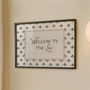 May include: A framed sign with the words "Welcome to the Loo" hangs on a cream-colored wall above blue tiles. The sign has a black frame and a white background with a black floral pattern. A round, white light fixture is to the right.
