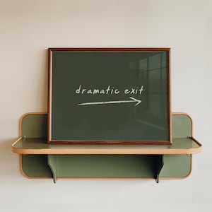 May include: A framed chalkboard with the words "dramatic exit" and an arrow pointing right. The frame is dark brown, and the chalkboard is mounted on a green and tan shelf against a cream-colored wall.
