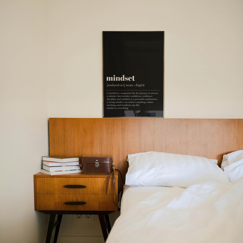 K&ouml;nnte beinhalten: Schlafzimmer mit einem Nachttisch aus Holz, B&uuml;chern und einer kleinen braunen Ledertasche. Ein schwarzer gerahmter Druck an der Wand zeigt das Wort "Mindset" und seine Definition. Ein wei&szlig;es Bett und ein h&ouml;lzernes Kopfteil sind ebenfalls zu sehen.