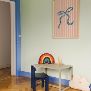 Pode incluir: Um quadro retangular com fundo de riscas verticais cor-de-rosa e brancas e uma ilustra&ccedil;&atilde;o central de um la&ccedil;o azul numa moldura de madeira clara, colocado sobre uma secret&aacute;ria cinzenta, uma cadeira azul-escura e duas almofadas.