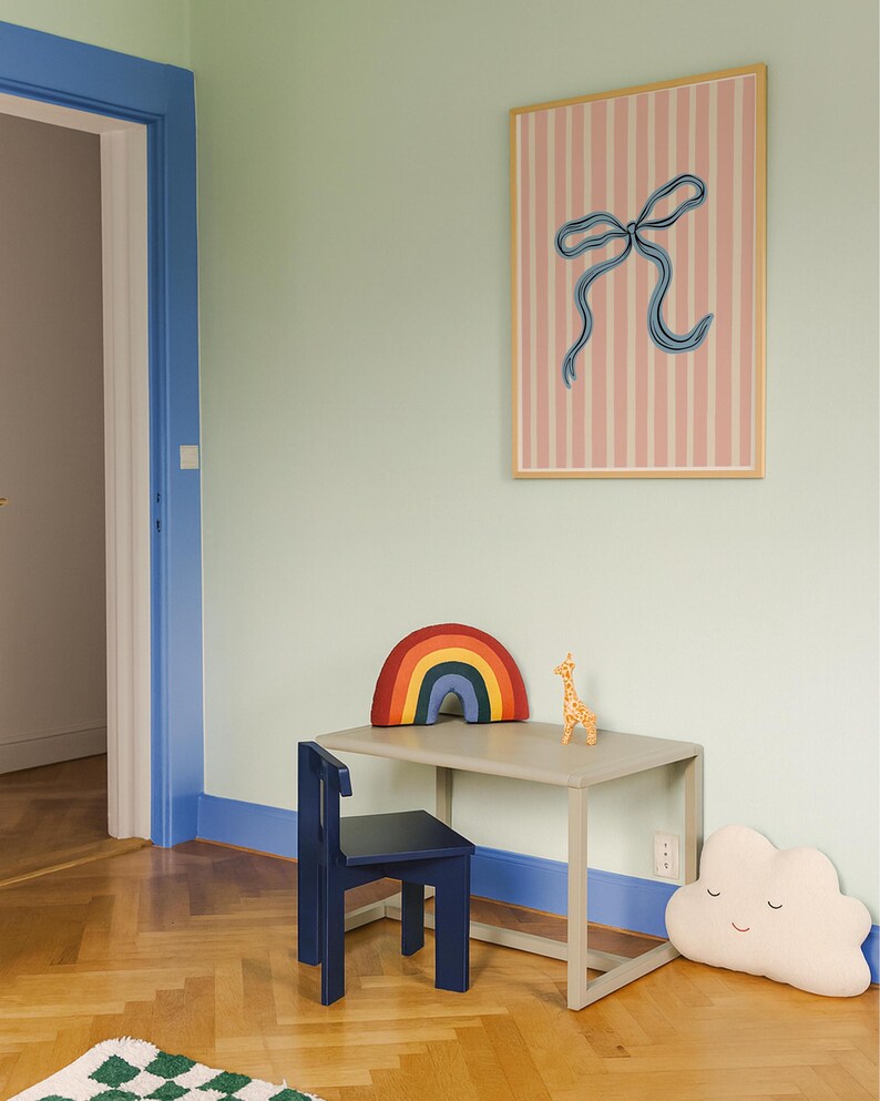 Pode incluir: Um quadro retangular com fundo de riscas verticais cor-de-rosa e brancas e uma ilustra&ccedil;&atilde;o central de um la&ccedil;o azul numa moldura de madeira clara, colocado sobre uma secret&aacute;ria cinzenta, uma cadeira azul-escura e duas almofadas.