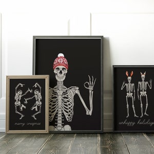 Op de afbeelding: Drie ingelijste wandposters met skeletten op een zwarte achtergrond. De linker poster toont twee dansende skeletten met de tekst "merry creepmas". De middelste poster toont een skelet met een rode gebreide muts. De rechter poster toont twee skeletten met een gewei en de tekst "unhappy holidays".