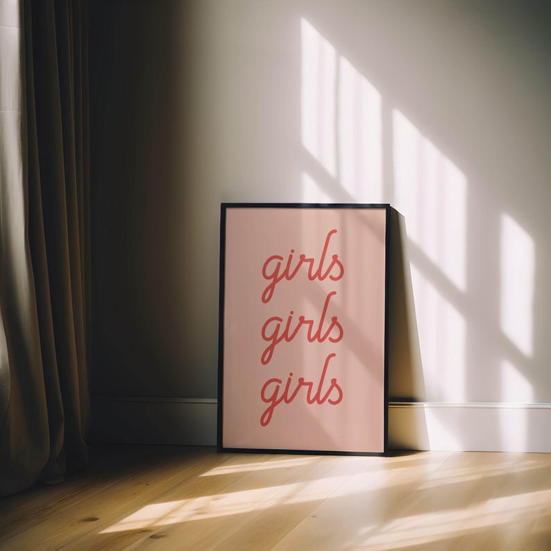May include: A framed pink poster with the word "girls" repeated three times in a cursive font. The poster is leaning against a white wall on a wooden floor. Sunlight casts shadows across the wall and floor.