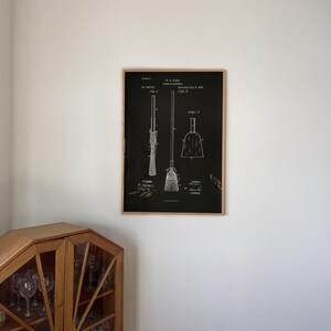 May include: A framed print featuring a broom patent design in black and white. The print shows detailed diagrams of a broom, dustpan, and related parts, with technical annotations and patent details. The frame is made of natural wood.