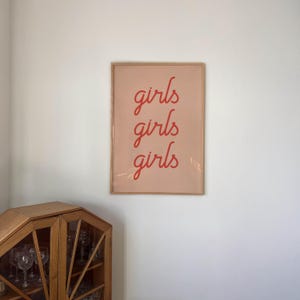 May include: A framed art print with the word "girls" repeated three times in a red script font. The background is a soft pink, and the frame is a light brown. The print is hanging on a white wall.