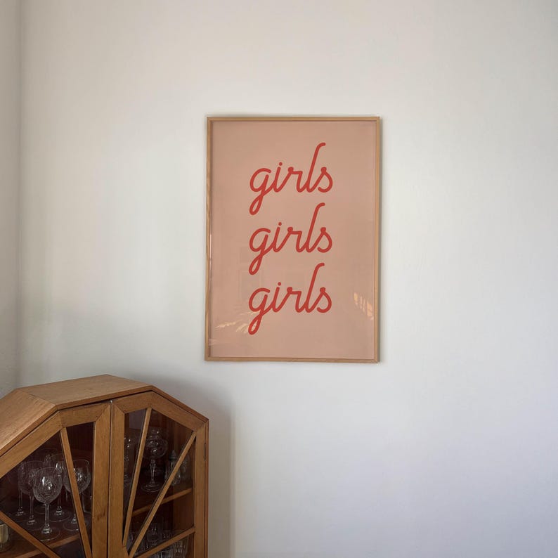 May include: A framed art print with the word "girls" repeated three times in a red script font. The background is a soft pink, and the frame is a light brown. The print is hanging on a white wall.