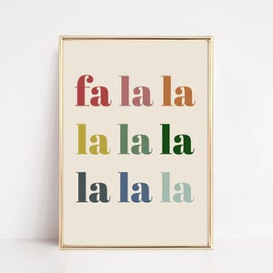 以下が含まれることがあります： カラフルなテキストで「fa la la」という言葉が重ねられた額入りのプリント。テキストは赤、ピンク、オレンジ、黄色、緑、青、ターコイズ色です。背景は薄いベージュ色です。